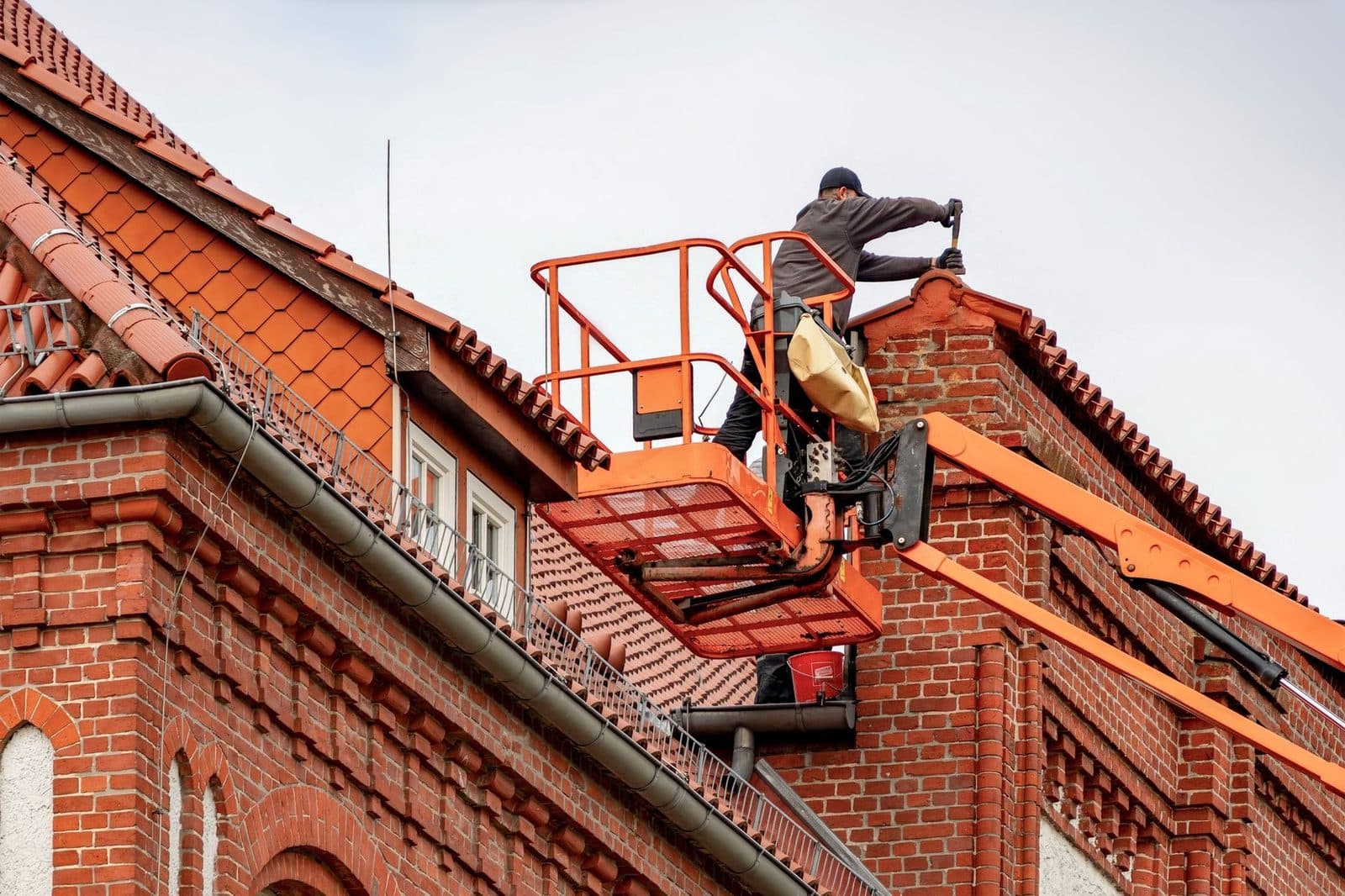 Ouvrier sur nacelle élévatrice orange travaillant sur la toiture en tuiles d'un immeuble en briques rouges