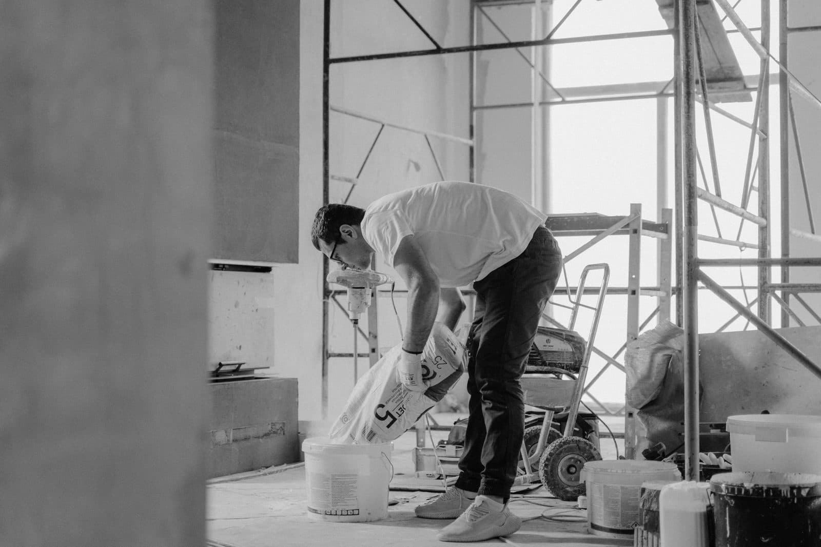 Ouvrier versant un sac d'enduit dans un seau sur un chantier de rénovation, échafaudage en arrière-plan