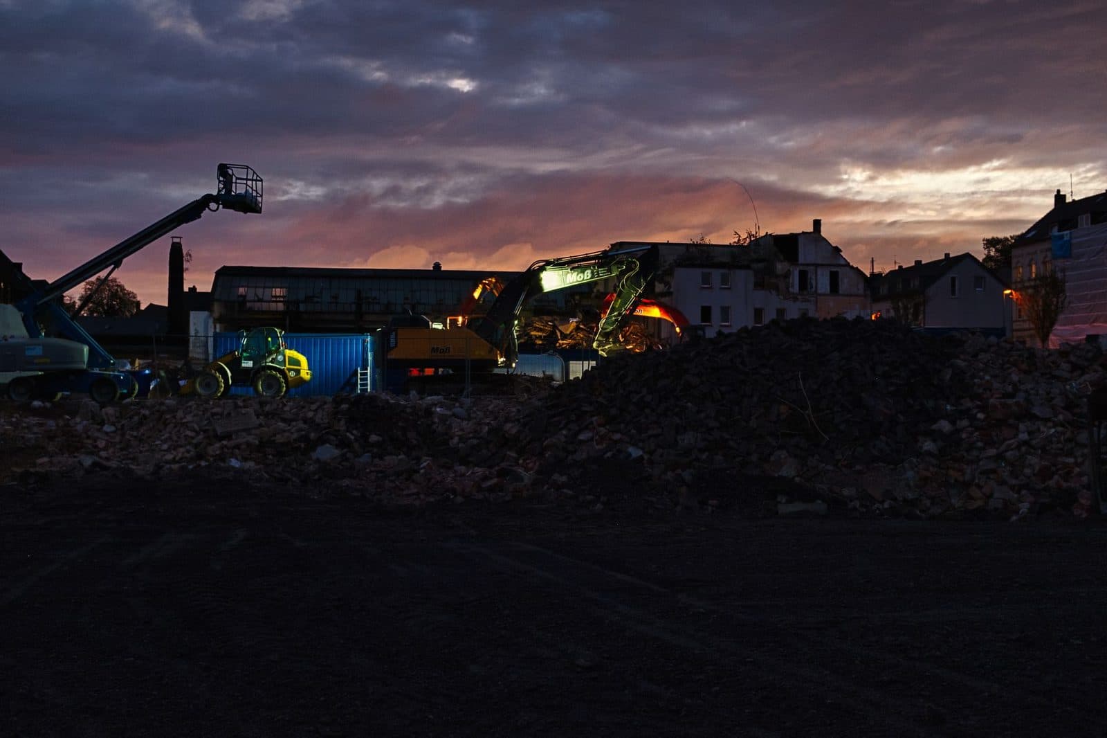 Engins de chantier et tas de gravats devant des bâtiments au crépuscule.