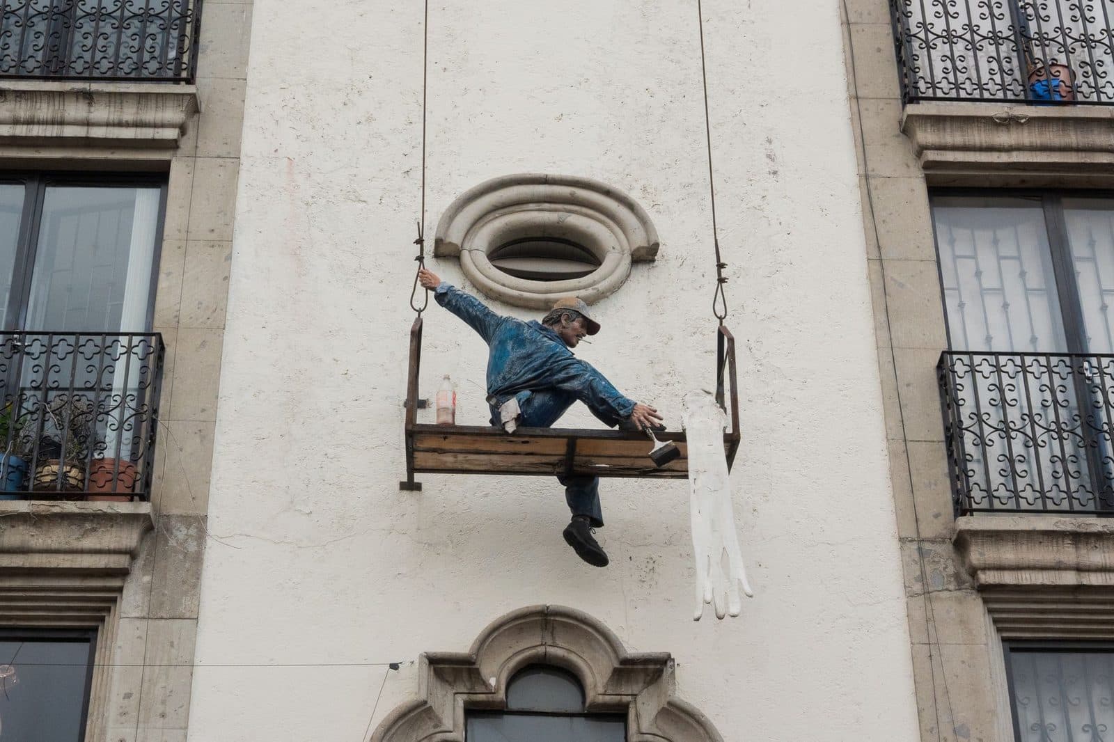 Sculpture d'un peintre en bâtiment sur une nacelle suspendue à la façade blanche d'un immeuble