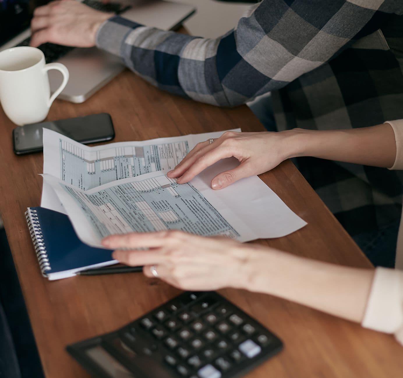 Deux personnes remplissent des formulaires administratifs sur une table en bois avec calculatrice et ordinateur portable