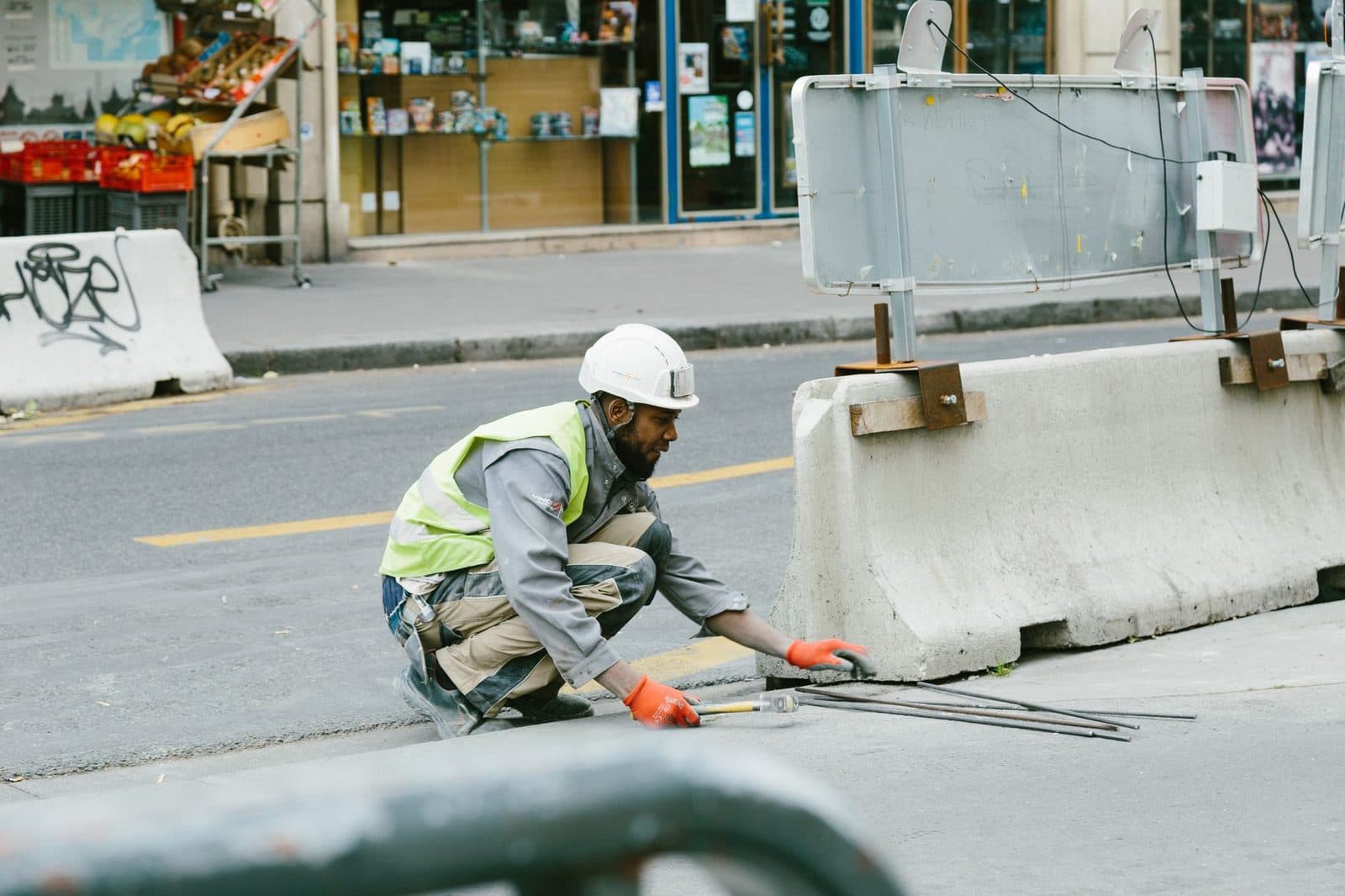 Ouvrier casqué en gilet jaune accroupi sur un chantier urbain près de blocs de béton et de tiges métalliques