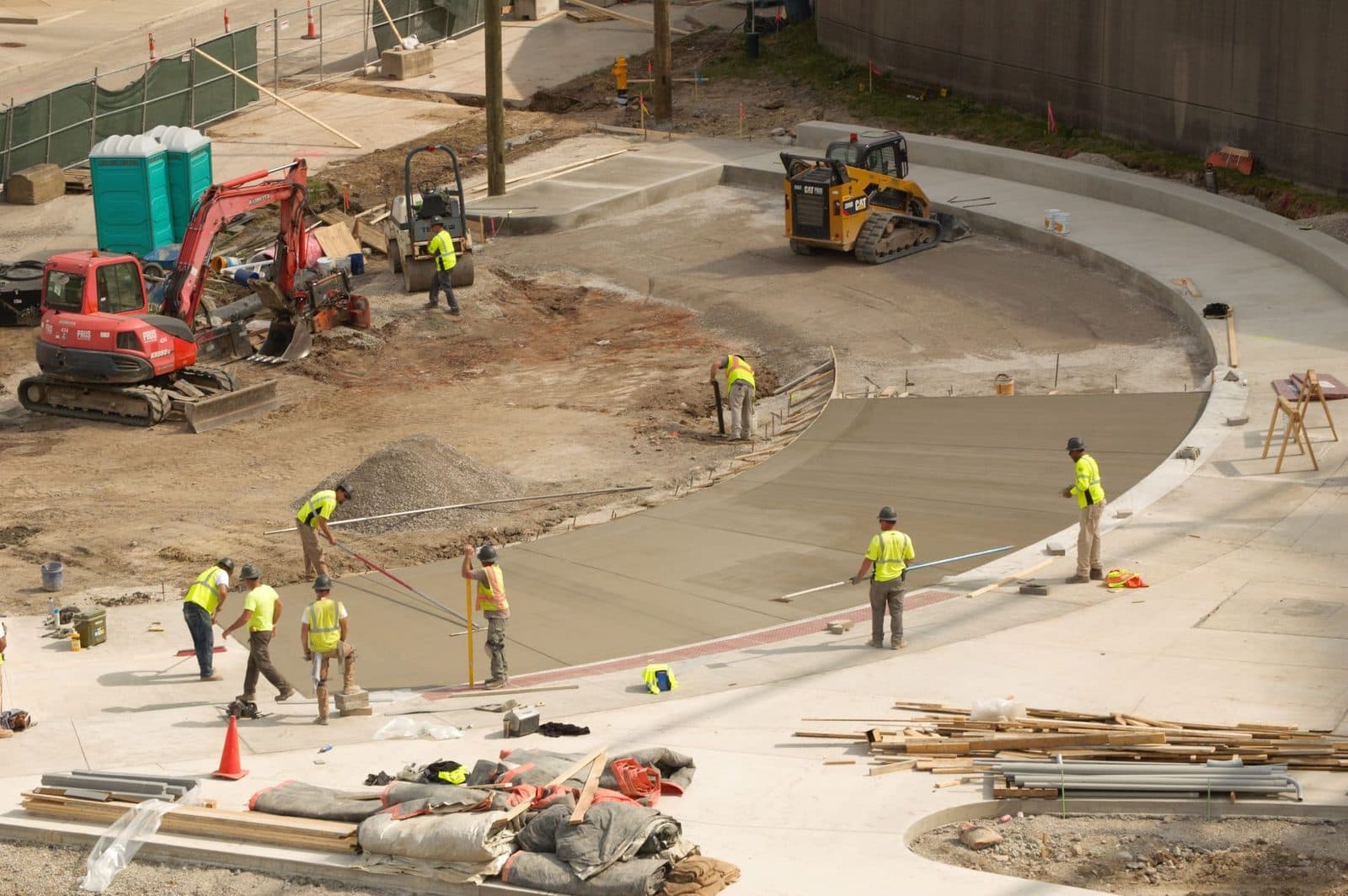 Ouvriers en gilets fluo lissant une dalle de béton sur un chantier, avec pelleteuse et chargeuse compacte