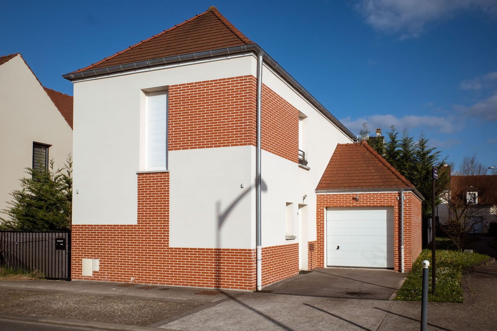 Maison individuelle neuve en briques rouges et enduit blanc avec garage, en bord de rue sous un ciel bleu