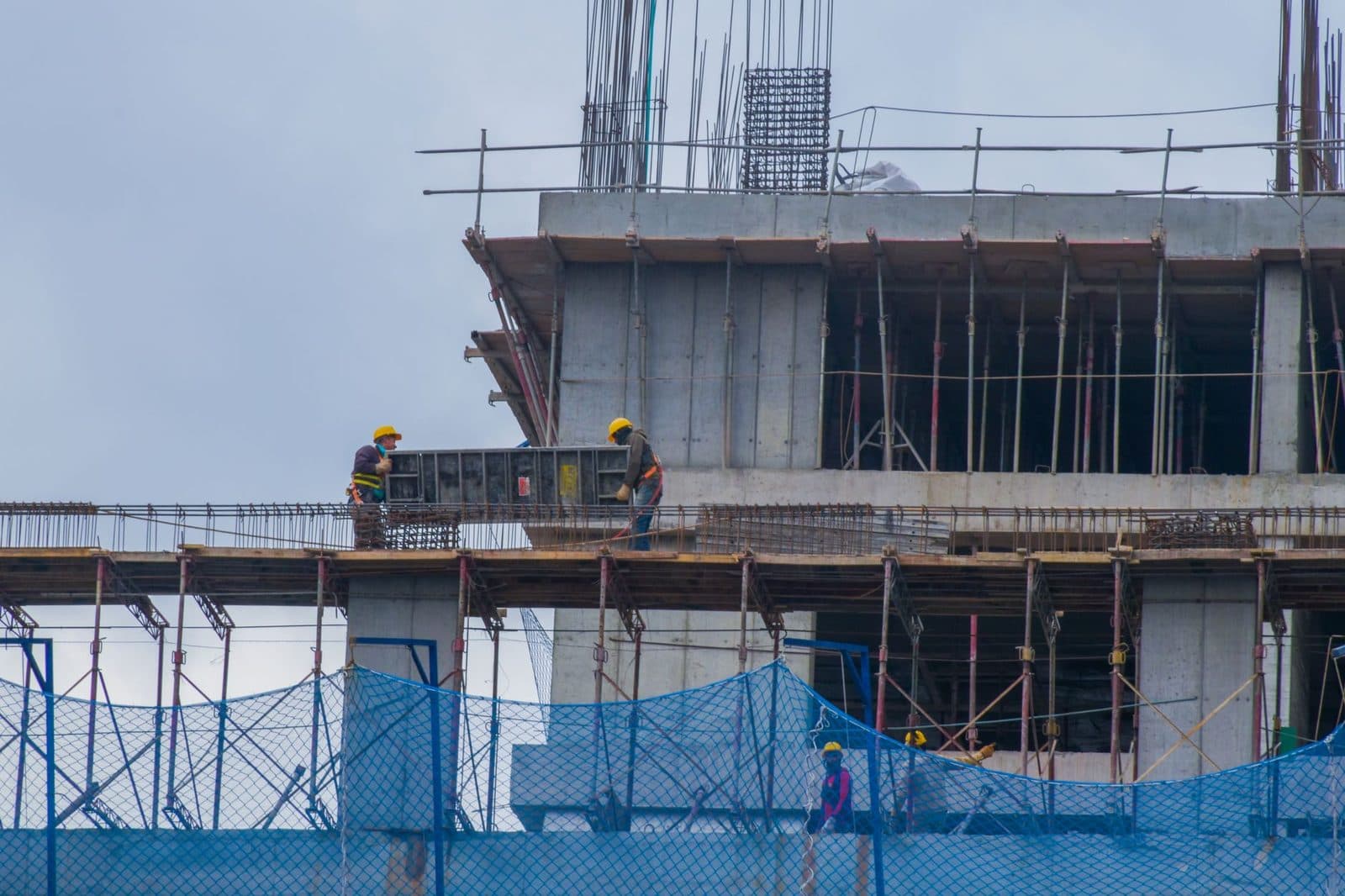 Ouvriers casqués sur un immeuble en construction en béton armé, entouré d'échafaudages et de filets bleus