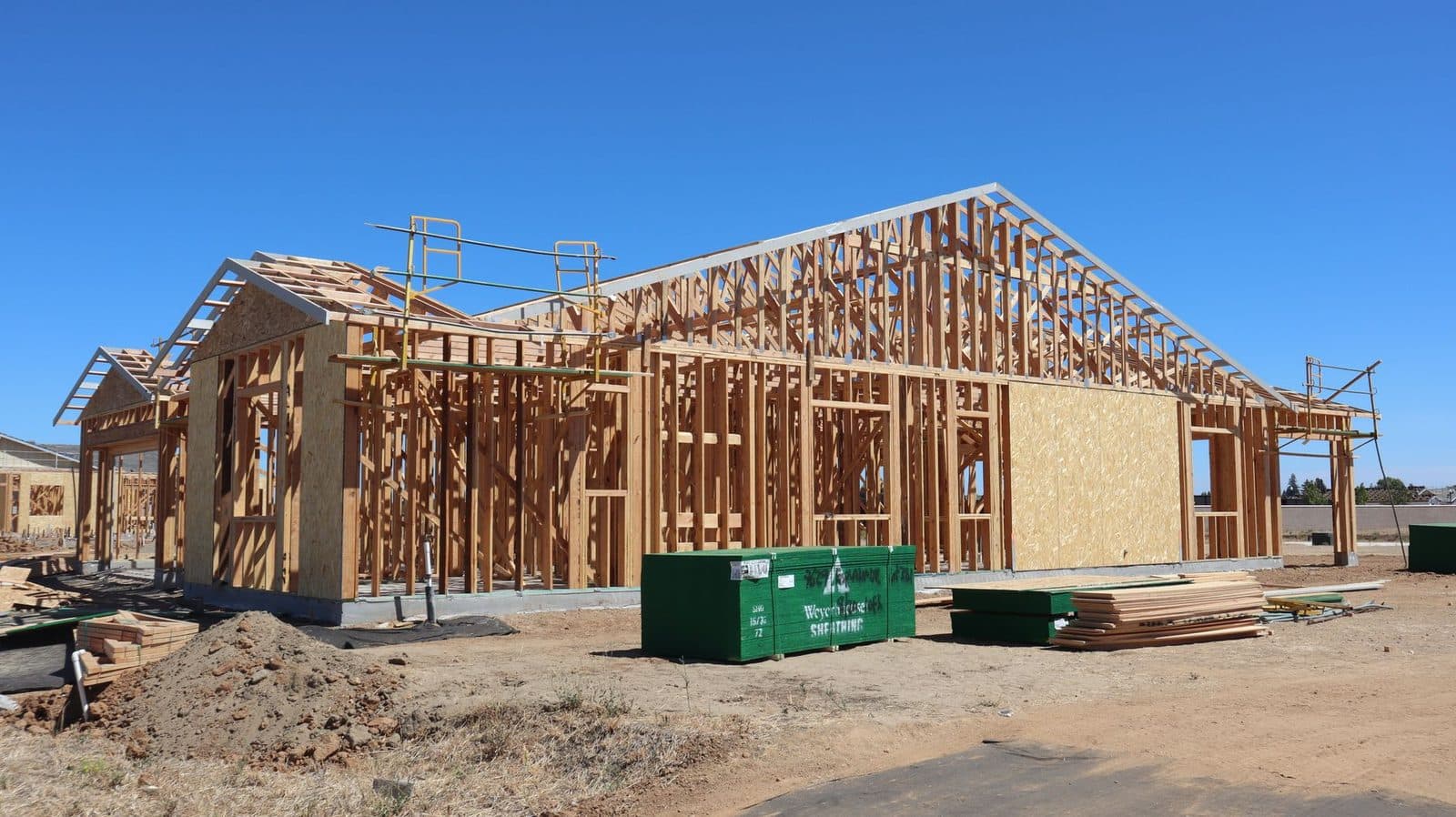 Maison en construction avec ossature en bois, benne verte et piles de planches sur le chantier