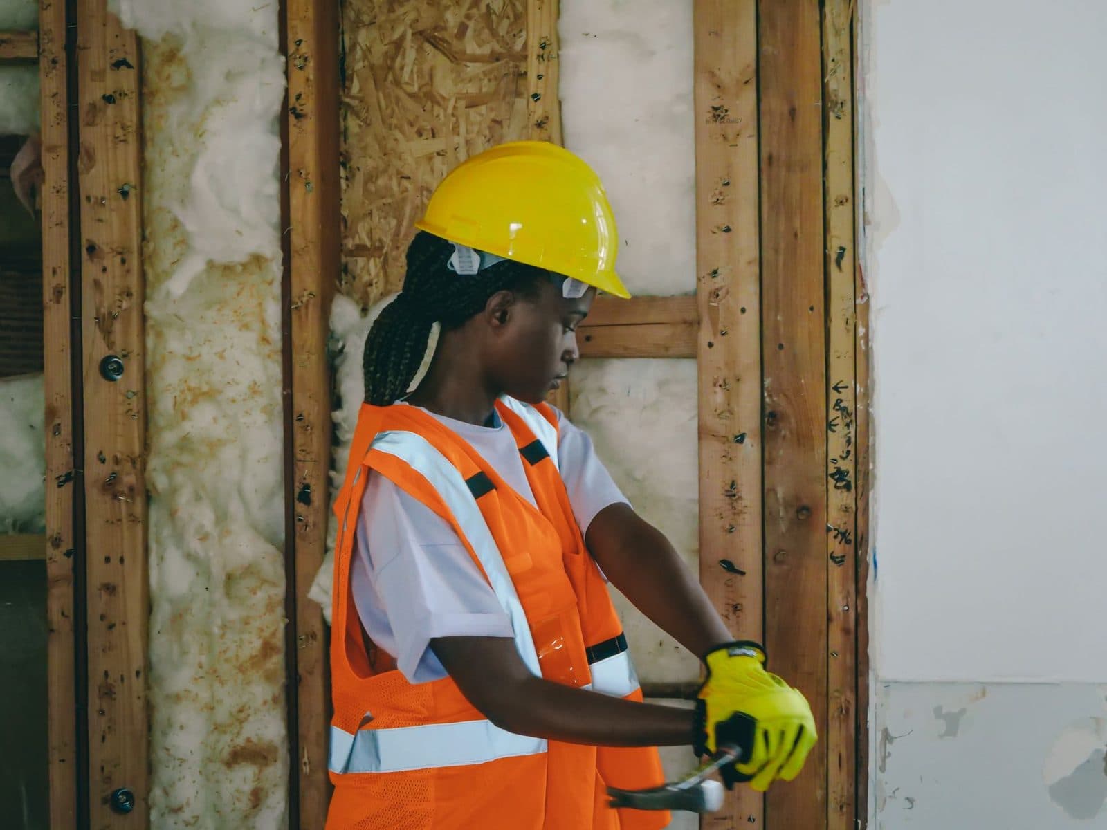 Ouvrière en casque jaune et gilet orange travaillant devant un mur ouvert avec ossature bois et isolation