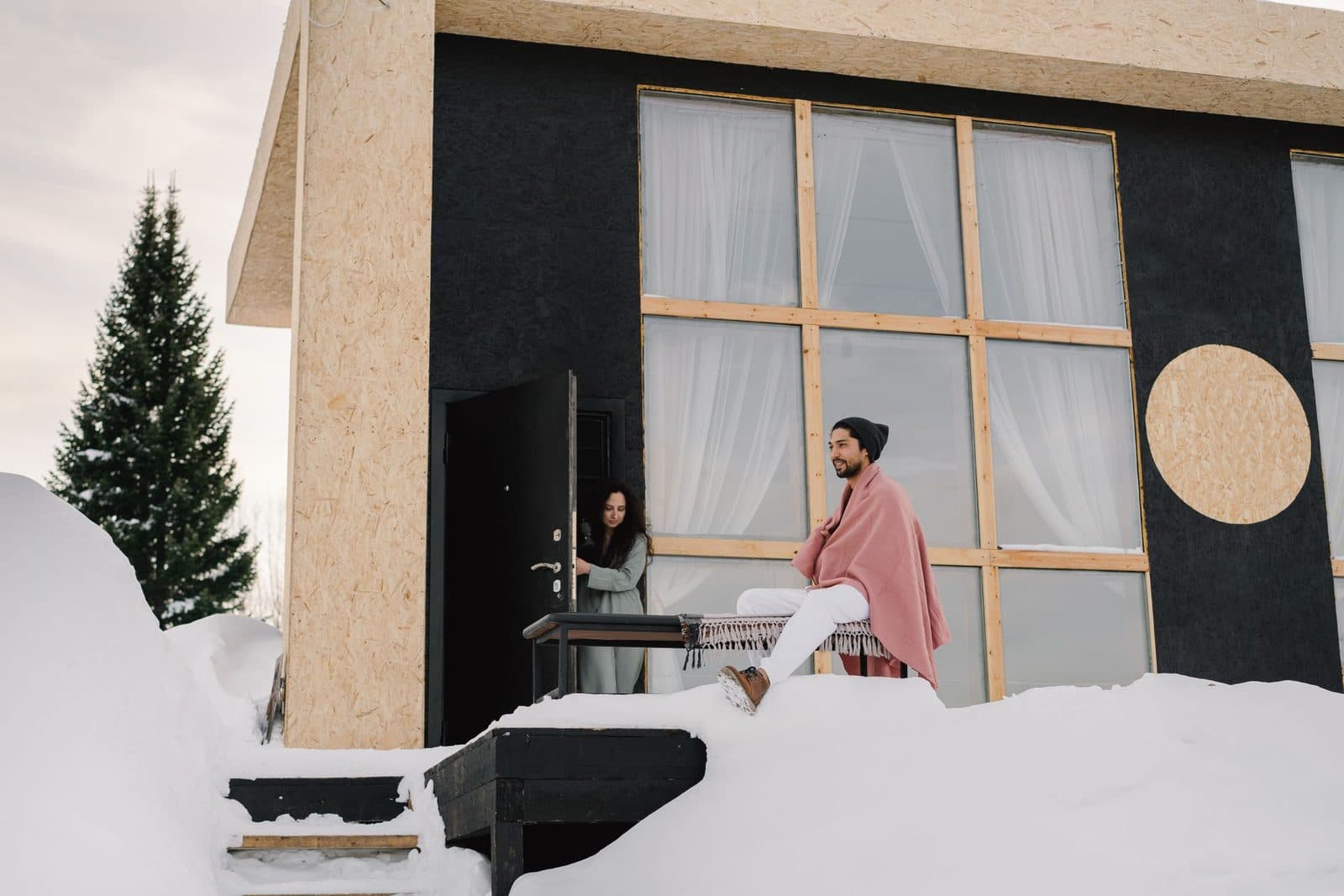 Couple devant une maison moderne en panneaux OSB avec grandes baies vitrées, sous la neige en hiver