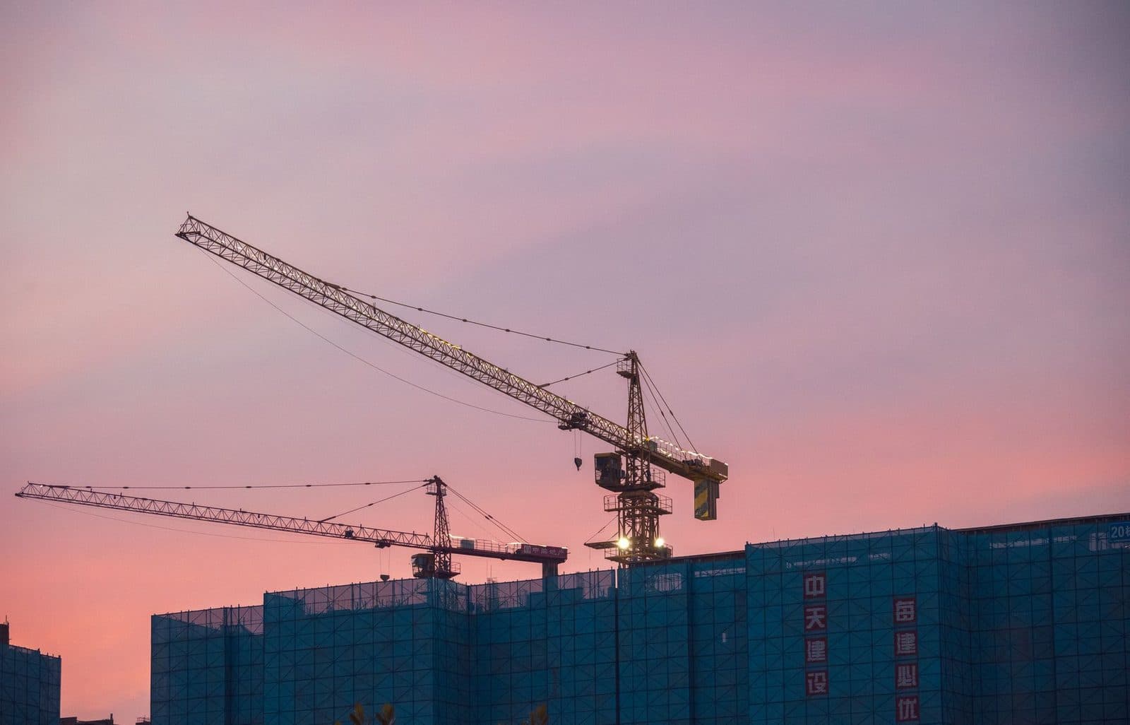 Deux grues à tour au-dessus d'un bâtiment en construction entouré de filets bleus, au coucher du soleil