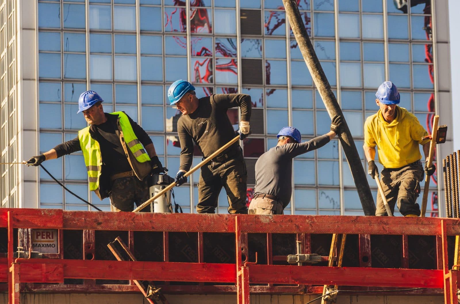 Quatre ouvriers en casque bleu travaillent sur un coffrage rouge d'un chantier, devant un immeuble vitré
