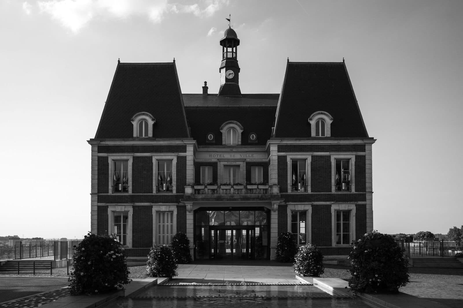 Façade d'un hôtel de ville en brique avec clocheton et horloge, massifs fleuris et bassin au premier plan