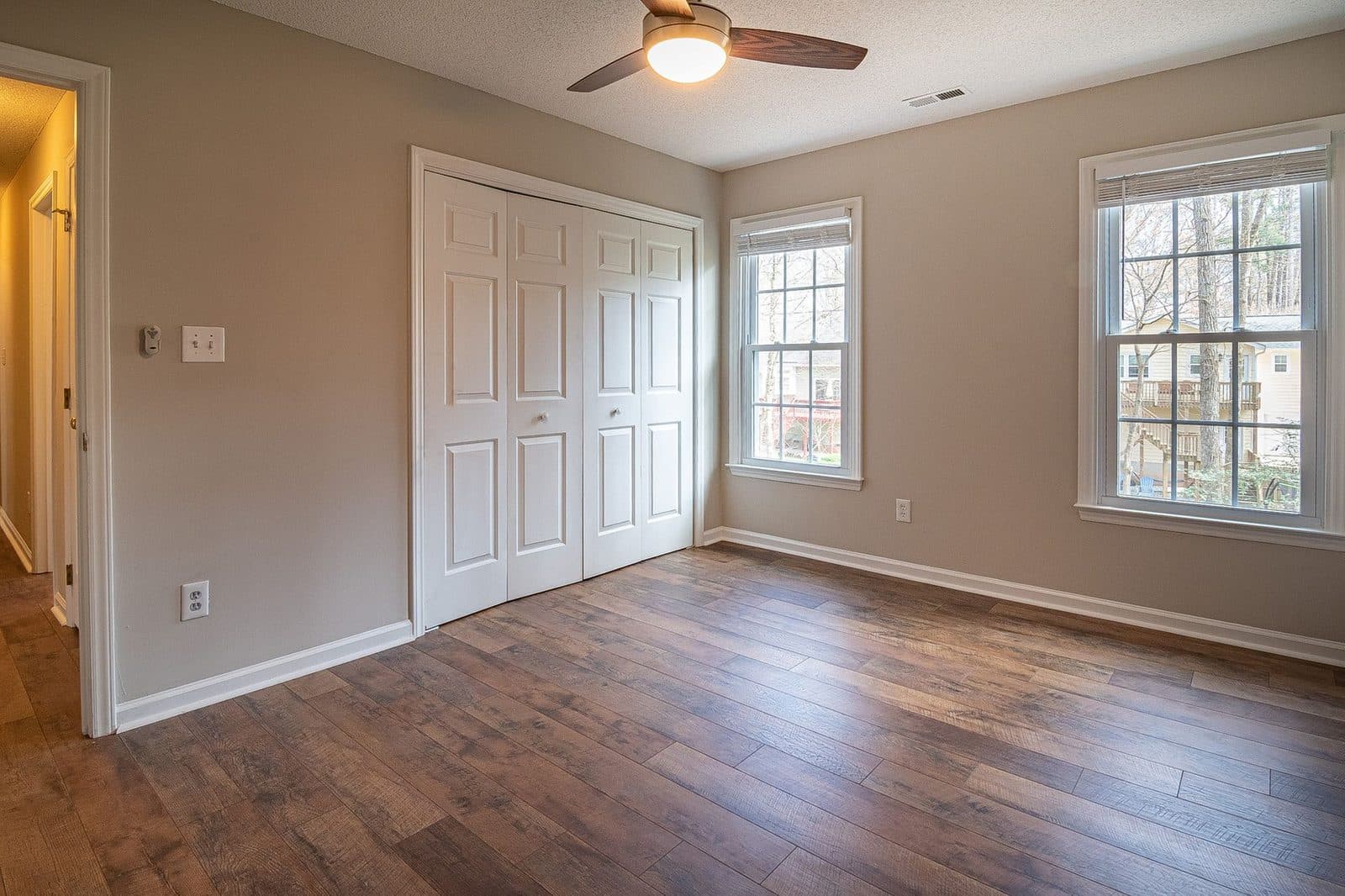 Pièce vide rénovée avec parquet, murs beiges, placard blanc, deux fenêtres et ventilateur de plafond