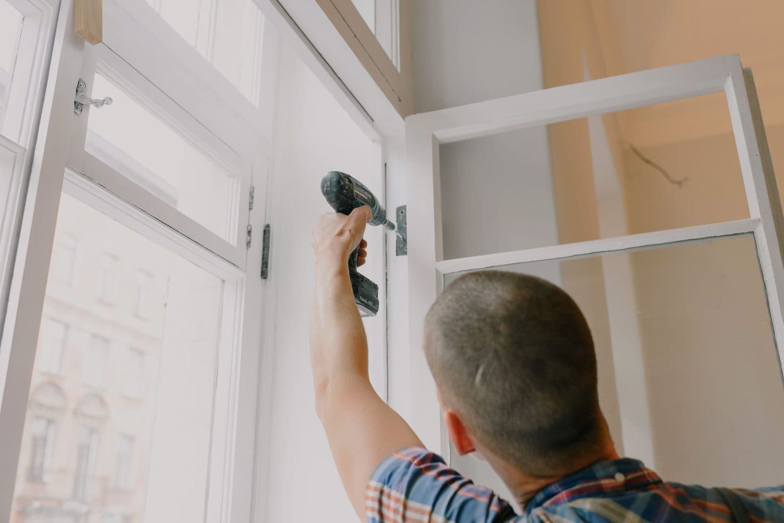 Homme en chemise à carreaux vissant un cadre de fenêtre blanc avec une perceuse lors de travaux de rénovation
