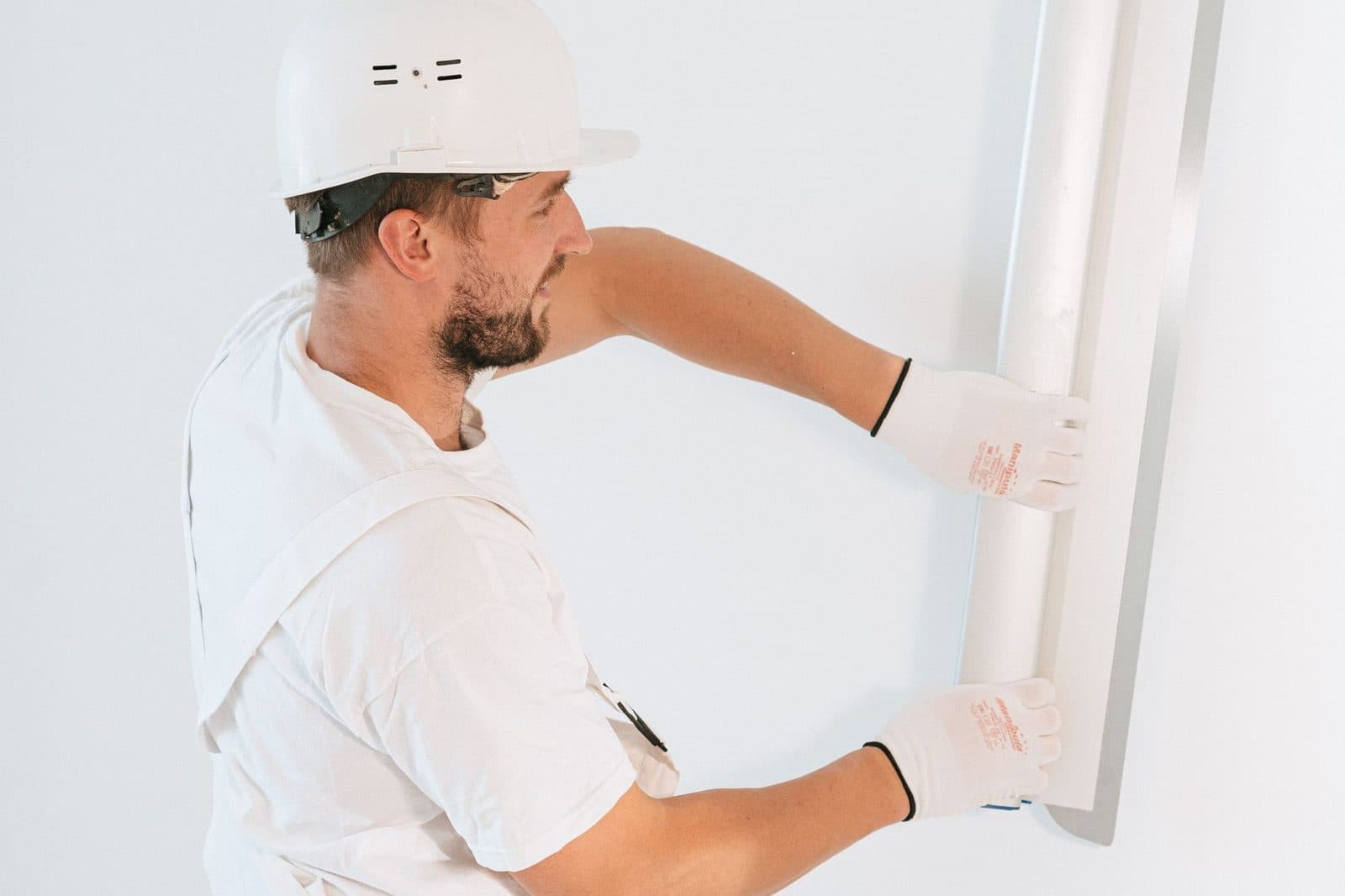 Ouvrier en casque et gants blancs posant un profilé métallique sur un mur blanc lors de travaux intérieurs