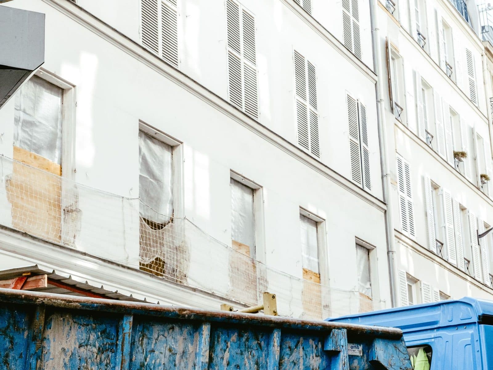 Immeuble haussmannien en rénovation avec fenêtres bâchées, filet de chantier et bennes bleues dans la rue