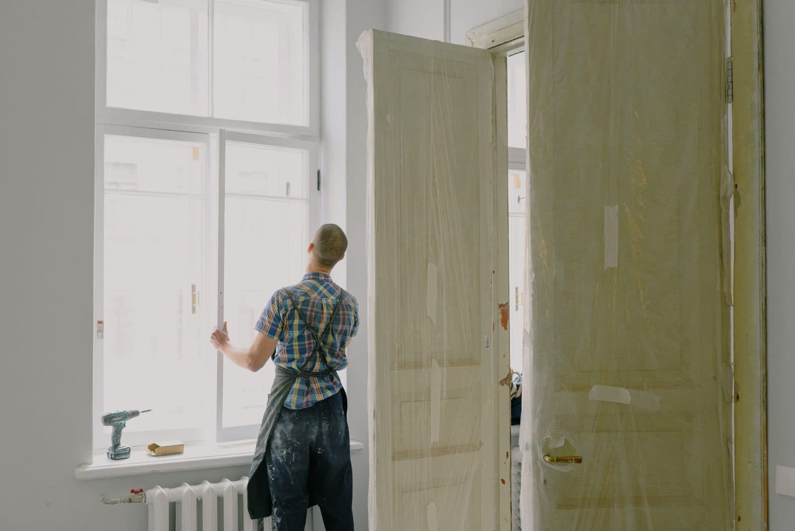 Ouvrier en salopette installant une fenêtre dans une pièce en travaux, portes protégées par du film plastique